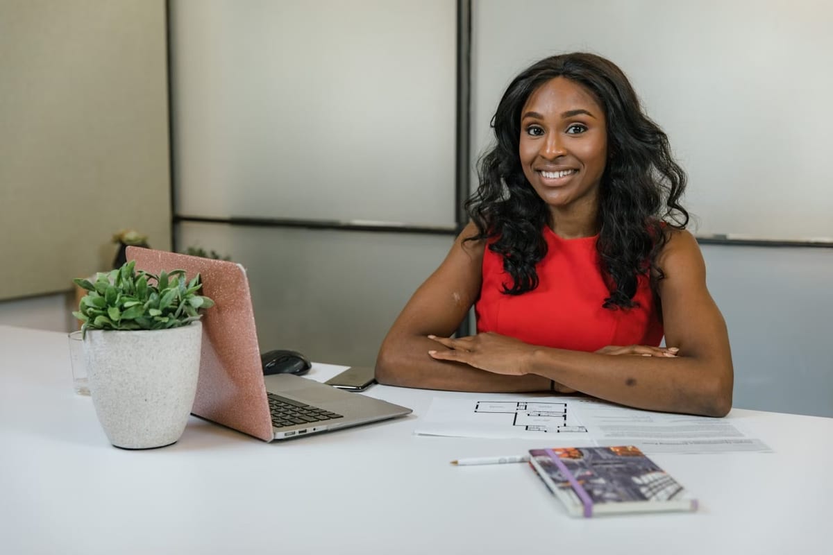 Empresária de blusa vermelha sorrindo à mesa de trabalho, com notebook, planta e planta baixa de projeto