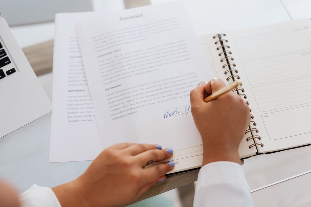 Pessoa assinando um contrato sobre uma mesa, com notebook e agenda ao lado
