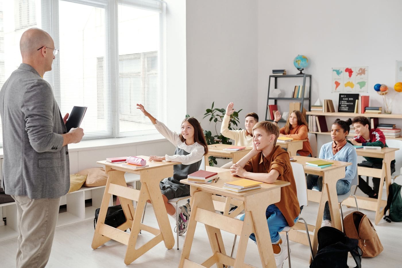 Professor e alunos em sala de aula, com estudantes levantando a mão para participar