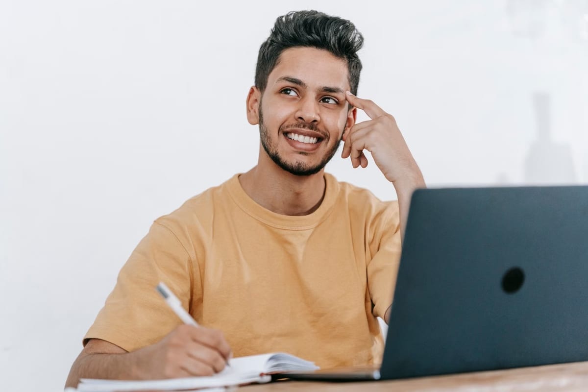 Homem sorrindo e pensativo, com a mão na testa, diante de um notebook e um caderno