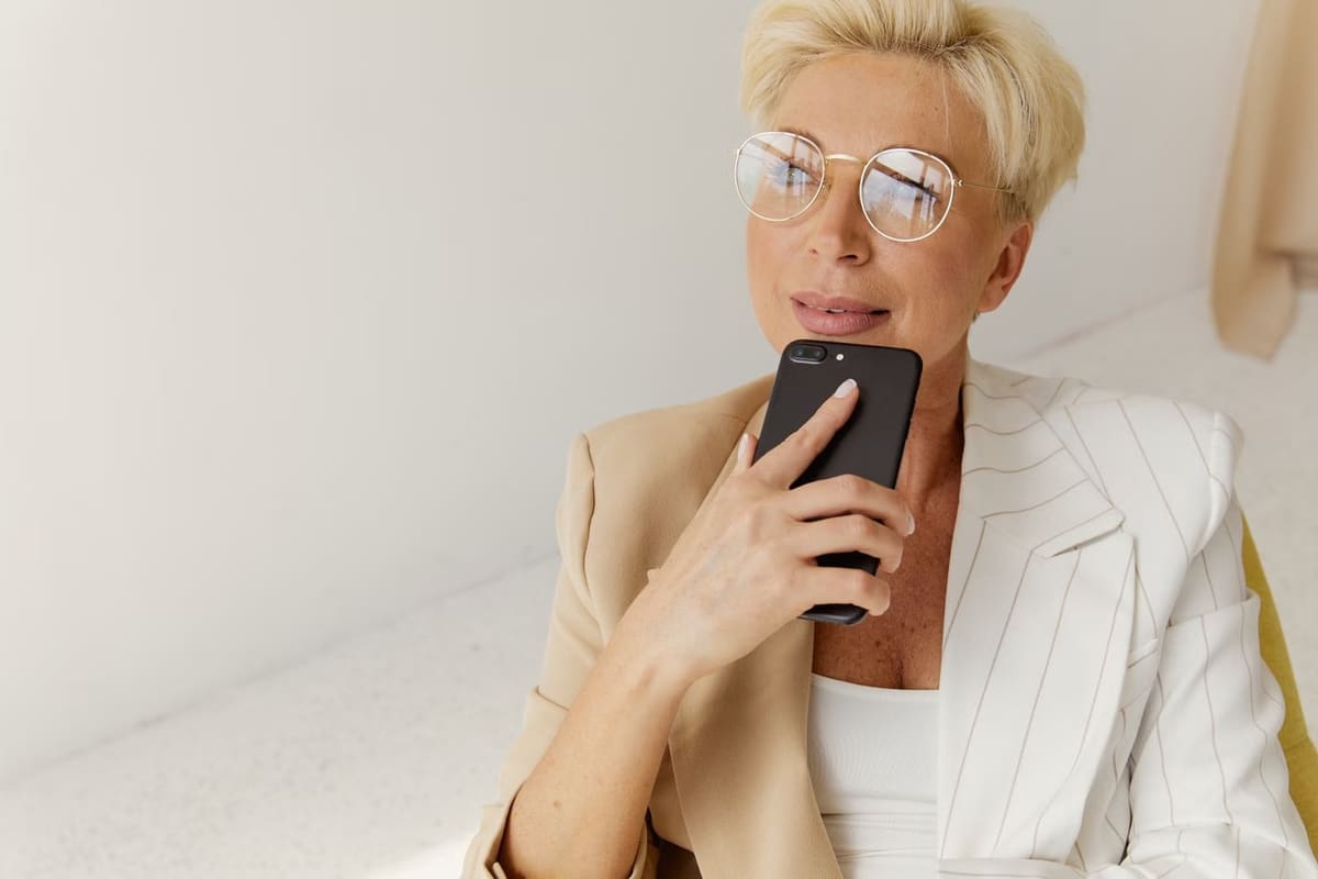 Mulher de blazer claro segurando o celular perto do rosto, pensativa, como quem espera uma resposta